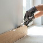 Mains gantées utilisant un outil pour retirer une plinthe en bois d'un mur gris, avec des morceaux de colle visibles.